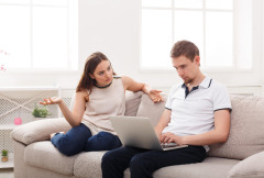 Image of a couple siting on a couch, the husband typing on a computer and the wife looking confused.